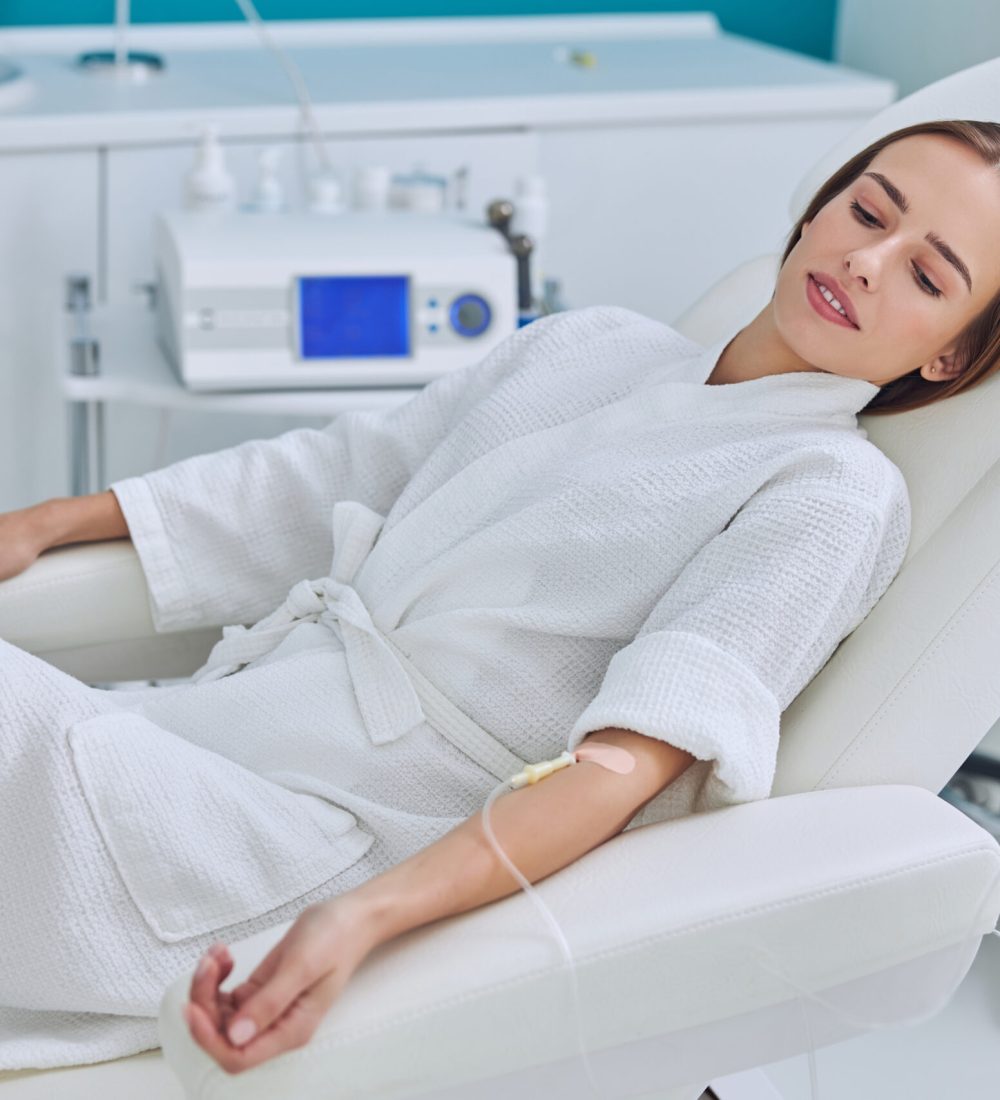 Waist up side view portrait of young pretty lady in white bathrobe lying on medicine armchair while receiving vitamin medical procedure in wellness center